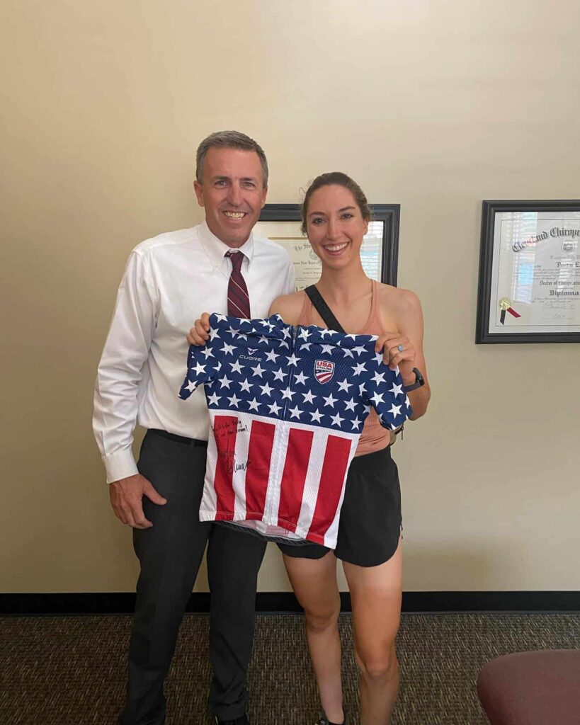 A man and a woman smile while holding up a USA Cycling jersey designed like the American flag in a chiropractic office.