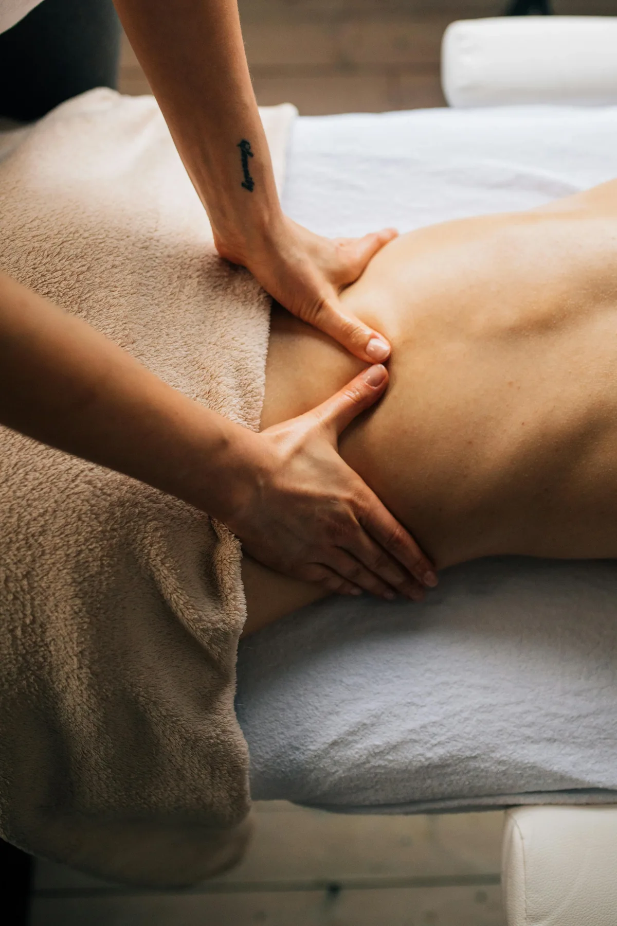 Close-up of a masseuse's hands massaging a client's lower back and upper gluteal area. The client is lying face down on a white massage table, partially covered by a beige towel. The masseuse's right forearm shows a small tattoo.