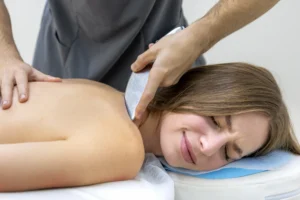 A woman lies on her stomach, eyes closed and face contorted in pain, as a therapist uses a curved metallic tool on her neck and shoulder.
