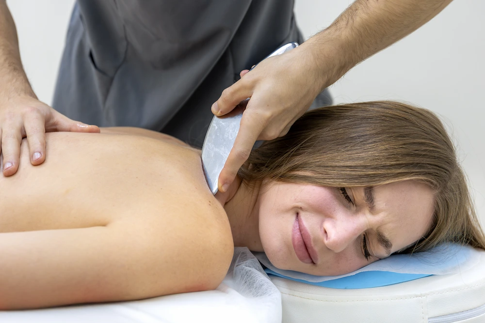 A woman lies on her stomach, eyes closed and face contorted in pain, as a therapist uses a curved metallic tool on her neck and shoulder.